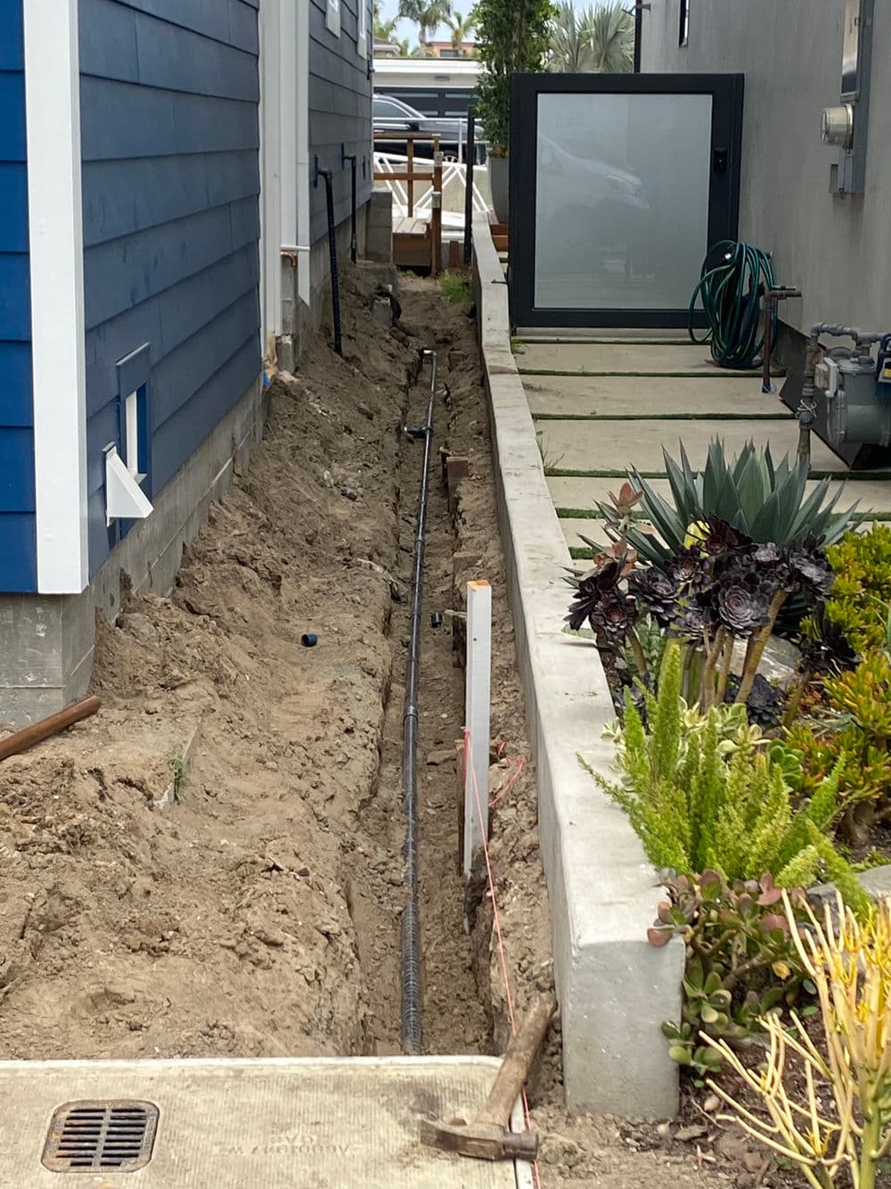Excavated trench for plumbing installation beside a blue house with landscaping.