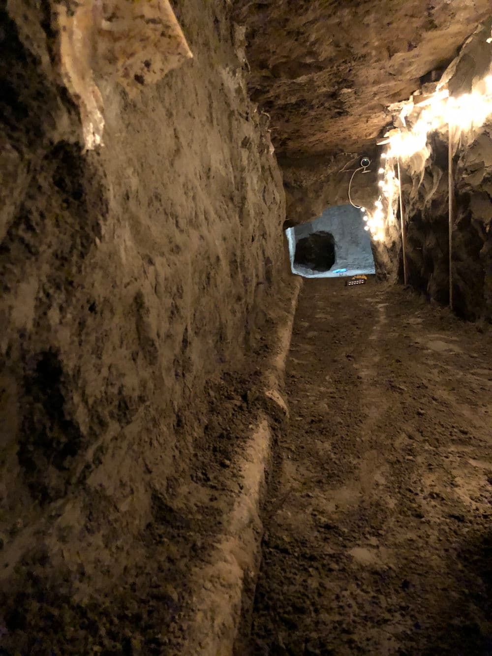 Narrow underground tunnel with illuminated walls leading to an opening in the distance.