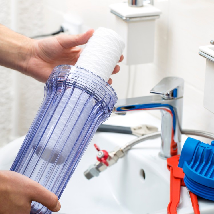 Hand holding a clear water filter cartridge above a sink, with plumbing tools and faucet visible, illustrating plumbing maintenance services by Plumbing By Tony.