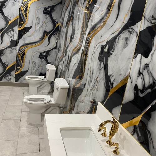 Modern restroom featuring elegant black and white marbled walls with gold accents, two white toilets, and a stylish sink with a vintage gold faucet.