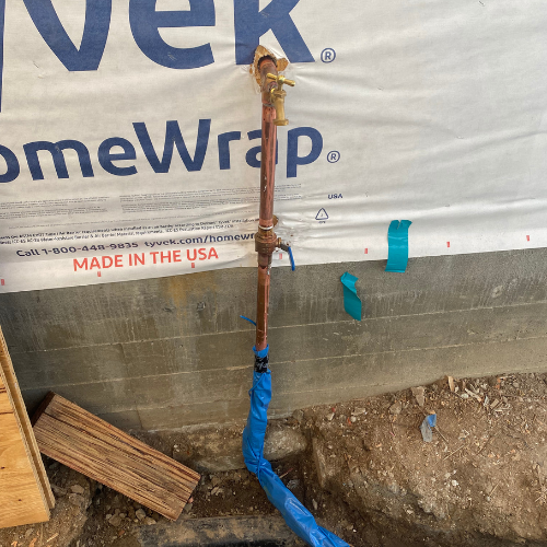 Plumbing By Tony Copper plumbing pipe with valve, wrapped in blue tape, against construction site backdrop with HomeWrap material, illustrating plumbing infrastructure for emergency repairs in Huntington Beach.